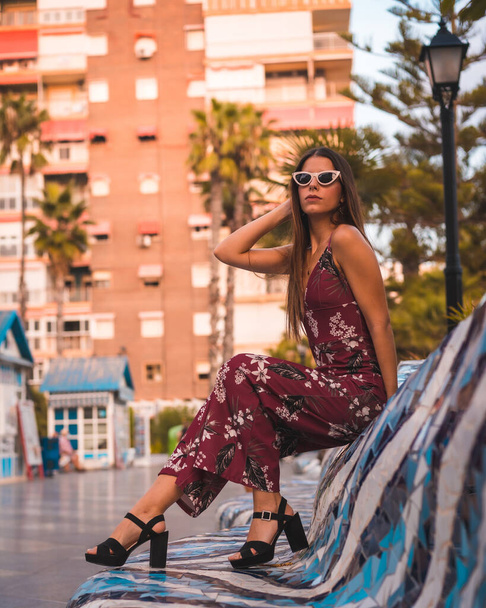Street Style, a young brunette in a maroon floral dress and modern white sunglasses enjoying the summer at the golden hour in the city - Photo, Image