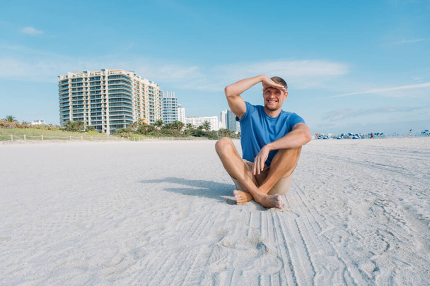 Un tipo caminando por la playa vacía de Miami. Feliz hombre guapo turista en ropa casual sentado en la playa de arena. Concepto de viajes y turismo. - Foto, imagen