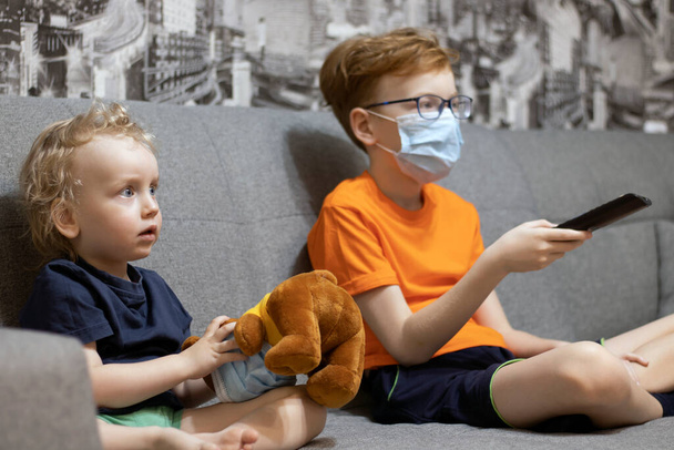 Children sitting on the couch in medical masks watch TV, the older brother switches the channel, holding the remote control in his hand. Quarantined. - Foto, Bild