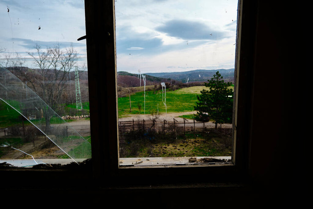 Kijkend naar buiten en het landbouwveld en het platteland achter de verlaten en bebouwde kom ramen. Glas van de ramen is gebroken. - Foto, afbeelding