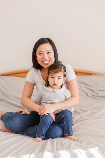 Asian Chinese woman hugging on bed with toddler girl at home. Girl daughter kid with mom together. Mother cuddling embracing with a baby daughter. Ethnic diversity.  - Photo, Image
