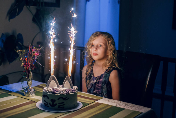 Cute funny blonde girl blowing candles on birthday cake in dark room. Happy birthday party celebration. Making a wish while blowing candles tradition. Child celebrating birthday at home. - Photo, Image