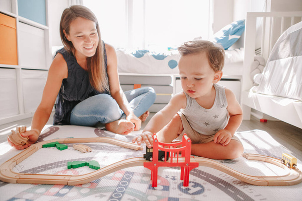 自宅で床に車の木製の鉄道で遊んで母親と幼児の少年。初期の教育開発。子供たちは鉄道道路を構築し、教育用おもちゃの列車、車を再生します。子供のためのレジャー活動.   - 写真・画像