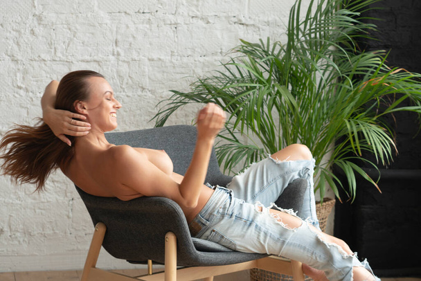 young beautiful woman posing nude, sitting in a chair in jeans - Photo, Image