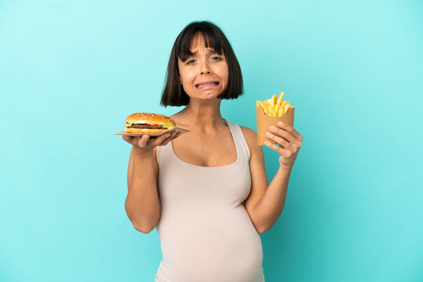 Молода вагітна жінка тримає бургер і смажені чіпси на ізольованому синьому фоні
 - Фото, зображення