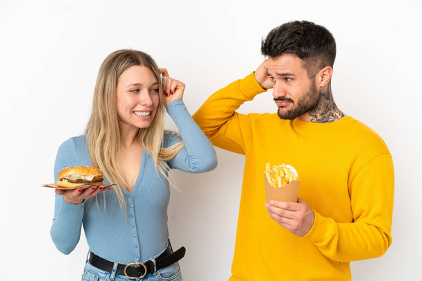 Couple holding hamburger and fried chips over isolated white background having doubts while scratching head - Foto, Bild