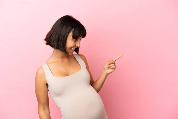 Junge schwangere Frau vor isoliertem rosa Hintergrund zeigt mit dem Finger zur Seite und präsentiert ein Produkt - Foto, Bild