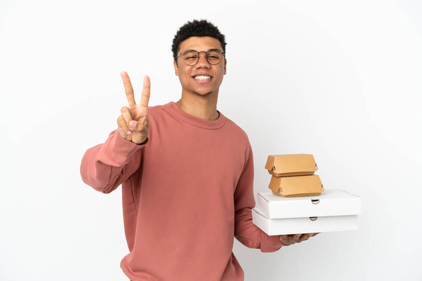 Beyaz arka planda izole edilmiş hamburger ve pizza tutan genç Afrikalı Amerikalı adam gülümsüyor ve zafer işareti gösteriyor. - Fotoğraf, Görsel