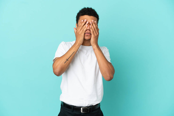 Νεαρός Αφροαμερικάνος απομονωμένος σε μπλε φόντο που καλύπτει τα μάτια με τα χέρια - Φωτογραφία, εικόνα