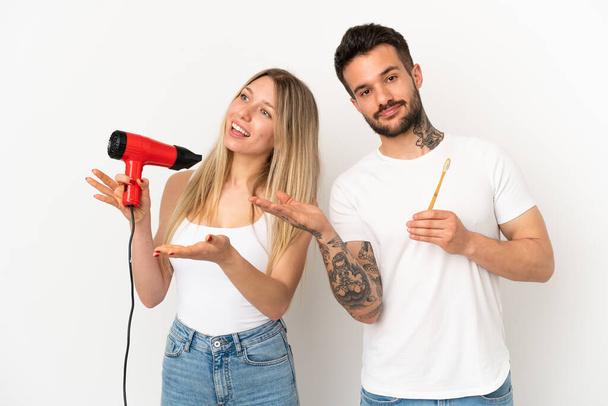 Couple holding a hairdryer and brushing teeth over isolated white background extending hands to the side for inviting to come - Φωτογραφία, εικόνα