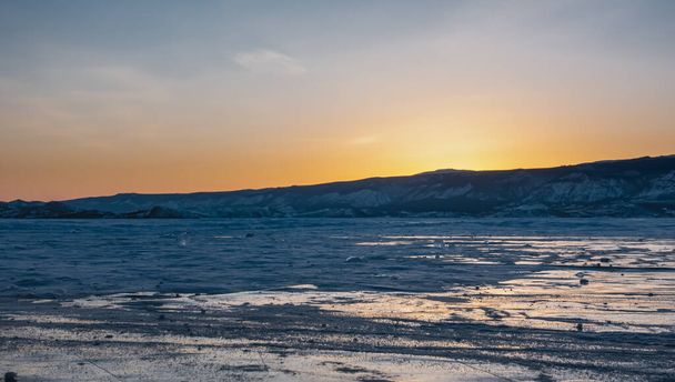 Zachód słońca nad zamarzniętym jeziorem. Niebo nad pasmem górskim jest koloru pomarańczowego. Na lodzie są plamy śniegu i odbicia zachodzącego słońca. Bajkał - Zdjęcie, obraz