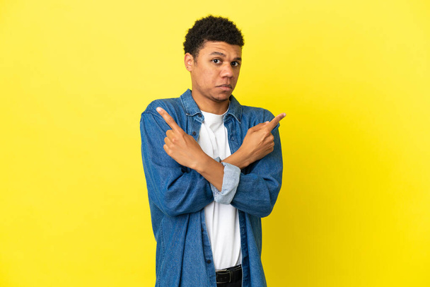 Young African American man isolated on yellow background pointing to the laterals having doubts - Фото, зображення