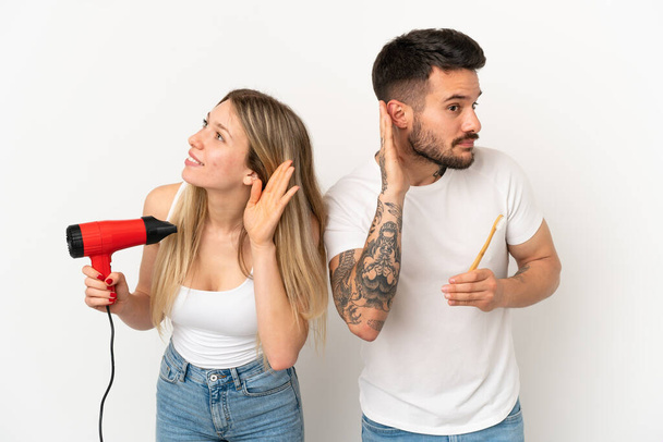 Couple holding a hairdryer and brushing teeth over isolated white background listening to something by putting hand on the ear - Photo, Image