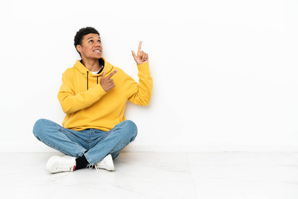 Молодой африканский американец сидит на полу изолированный на белом фоне указывая указательным пальцем отличная идея - Фото, изображение