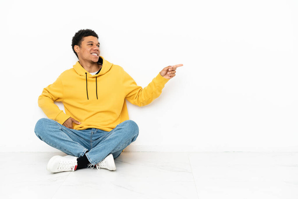 Genç Afro-Amerikalı bir adam beyaz arka planda izole bir şekilde oturuyor. Parmakla göstererek bir ürün sunuyor. - Fotoğraf, Görsel
