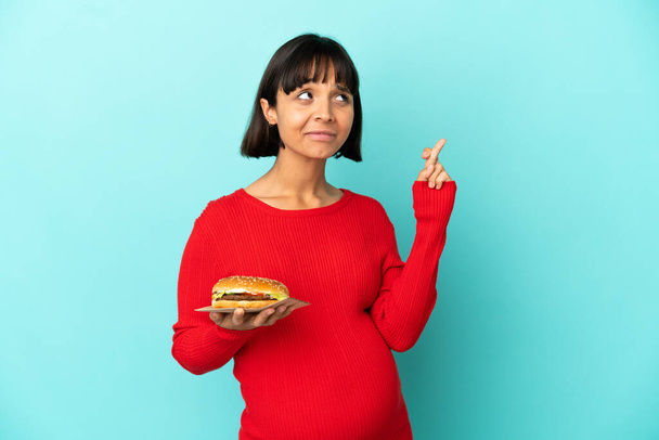 Молода вагітна жінка тримає бургер над ізольованим тлом з пальцями перетинаючи і бажаючи кращого
 - Фото, зображення