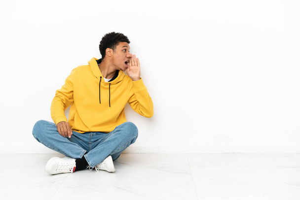 Mladý Afroameričan sedí na podlaze izolovaný na bílém pozadí křičí s ústy dokořán otevřenými bočním - Fotografie, Obrázek
