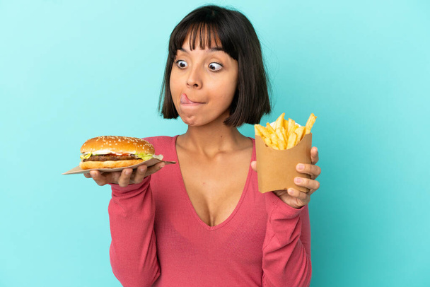 jonge brunette vrouw met hamburger en gebakken chips over geïsoleerde blauwe achtergrond - Foto, afbeelding