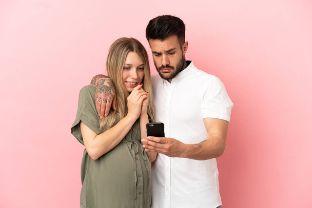 Donna incinta e uomo su sfondo rosa isolato leggendo un messaggio con il cellulare - Foto, immagini