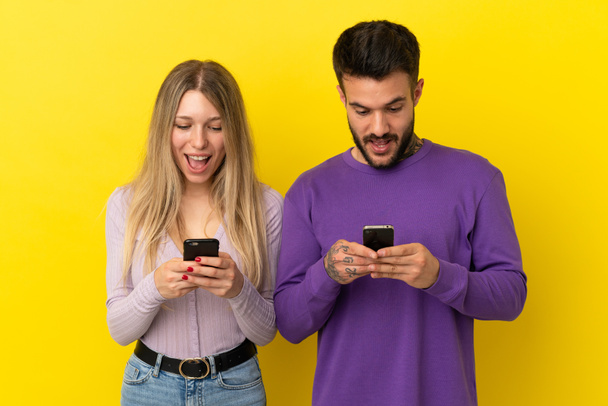 孤立した黄色の背景に若いカップルは驚いて、モバイルでメッセージや電子メールを送信します - 写真・画像