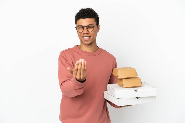 Giovane uomo afroamericano che tiene un hamburger e pizze isolate su sfondo bianco invitando a venire con mano. Felice che tu sia venuto - Foto, immagini