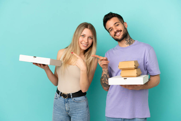 пара держит пиццу и бургеры на изолированном синем фоне указывая пальцем в сторону в боковом положении - Фото, изображение