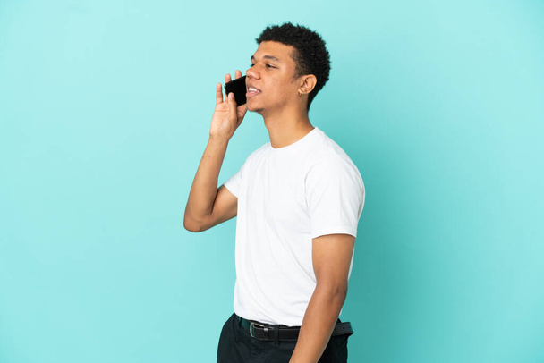 Young African American man isolated on blue background keeping a conversation with the mobile phone - Φωτογραφία, εικόνα