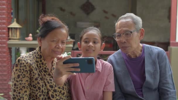 Ευτυχισμένο ασιατικό αξιολάτρευτο κορίτσι κάθεται δίπλα στους ηλικιωμένους παππούδες της και κρατώντας κινητό smartphone για να δείξει και να παρακολουθήσουν κοινωνικό χαρτοφυλάκιο βίντεο της μαζί στο σπίτι. Σχέση δεσμού στην οικογένεια. - Πλάνα, βίντεο