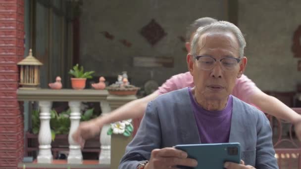 Vidám ázsiai csinos lány jön és megöleli idős nagyapja hátulról, és élvezze nézni a közösségi média videó mobil okostelefon együtt otthon. Kötődés a családban. - Felvétel, videó