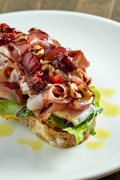 Huge sandwich with jamon crudo, brie cheese, mixed salad, grapes, sun-dried tomatoes and nuts on rye bread, served in a white plate on a wooden background. Delicious breakfast toast - Foto, Imagem