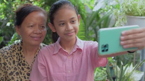 Cep telefonu tutan Asyalı sevimli kız yaşlı büyükannesiyle evdeki yeşil bahçede selfie çekmekten zevk alıyor. Sağlıklı bir yaşam tarzı. İki nesildir birlikteyiz.. - Video, Çekim