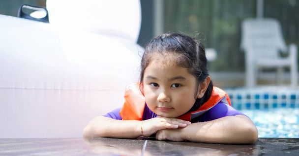 Asijské dítě hrající si v bazénu. Na sobě oranžovou záchrannou vestu, bradu opřenou o obě ruce na okraji bazénu. - Fotografie, Obrázek