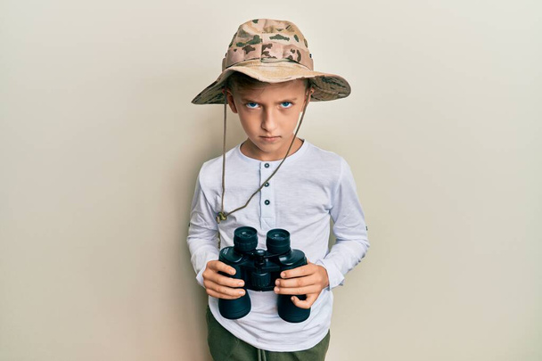 Un ragazzino caucasico che indossa un cappello da esploratore con in mano un binocolo scettico e nervoso, accigliato a causa di un problema. persona negativa.  - Foto, immagini