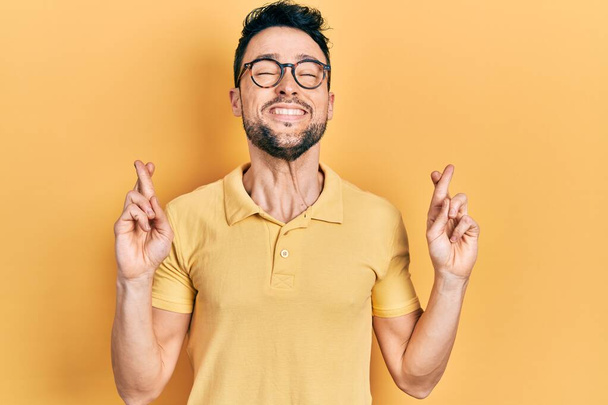 Young hispanic man wearing casual clothes and glasses gesturing finger crossed smiling with hope and eyes closed. luck and superstitious concept.  - Фото, изображение