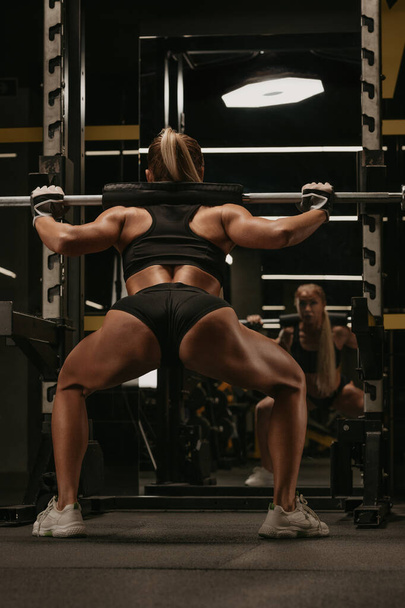Een foto van achteren van een sportieve vrouw met blond haar die hurkt met een lange halter in de buurt van het kraakrek in een sportschool. Een meisje doet een beentraining.. - Foto, afbeelding