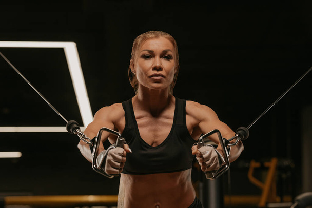 A close-up photo of a fit woman with blonde hair who is doing a chest workout on the cable machine in a gym. A girl is training her pectoral muscles. - Φωτογραφία, εικόνα