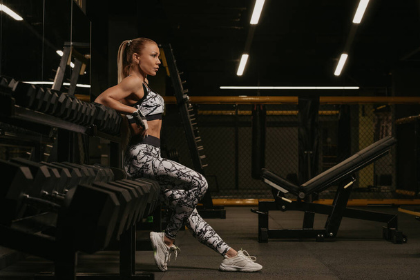 A photo from the side of a fit woman with blonde hair who is sitting on the row of dumbbells in a gym. A girl is relaxing after a workout. - Foto, Bild