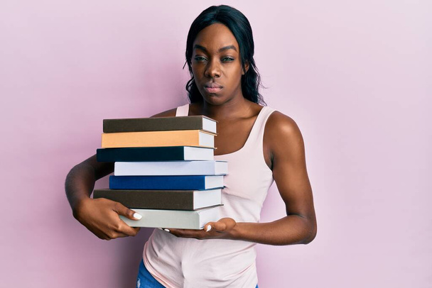 Mujer afroamericana joven sosteniendo libros escépticos y nerviosos, frunciendo el ceño molesto debido a un problema. persona negativa.  - Foto, imagen