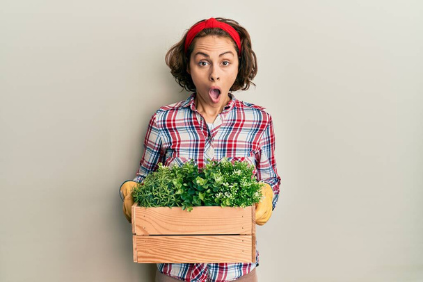 Young brunette woman wearing gardener clothes holding wooden plant pot afraid and shocked with surprise and amazed expression, fear and excited face.  - Fotoğraf, Görsel