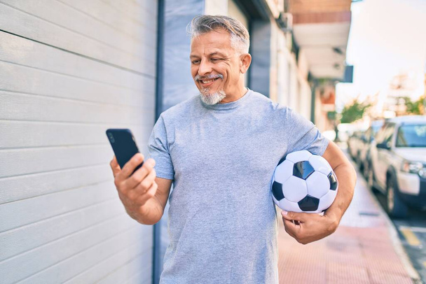 Middelbare leeftijd hispanic grijs-harige man met behulp van smartphone en het houden van voetbal in de stad. - Foto, afbeelding