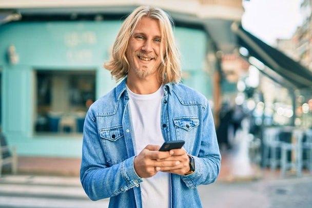 Jeune homme blond scandinave souriant heureux en utilisant un smartphone à la ville. - Photo, image