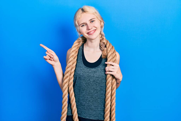 Красивая белая женщина с светлыми волосами тренирующаяся с боевой веревкой улыбаясь счастливо указывая рукой и пальцем в сторону  - Фото, изображение