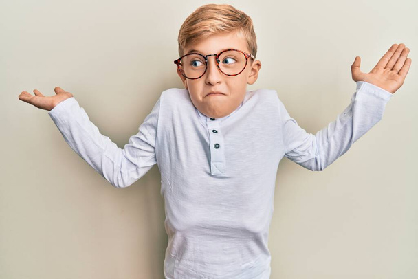 Kleiner kaukasischer Junge in lässiger Kleidung und Brille ahnungslos und verwirrt mit offenen Armen, keine Ahnung und zweifelhaftem Gesicht.  - Foto, Bild