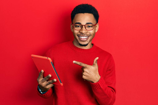 Jeune homme afro-américain utilisant un dispositif tactile souriant heureux pointant avec la main et le doigt  - Photo, image