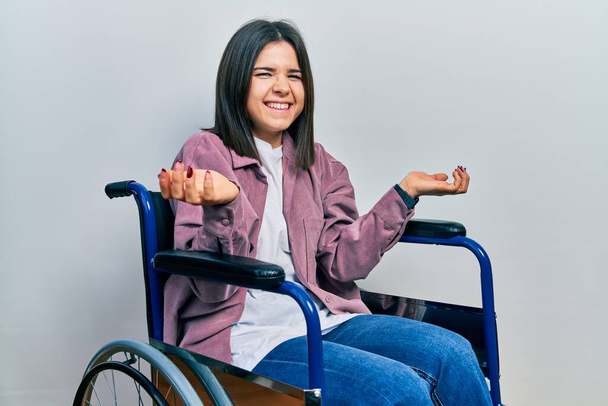 Young brunette woman sitting on wheelchair smiling cheerful with open arms as friendly welcome, positive and confident greetings  - Foto, Imagem