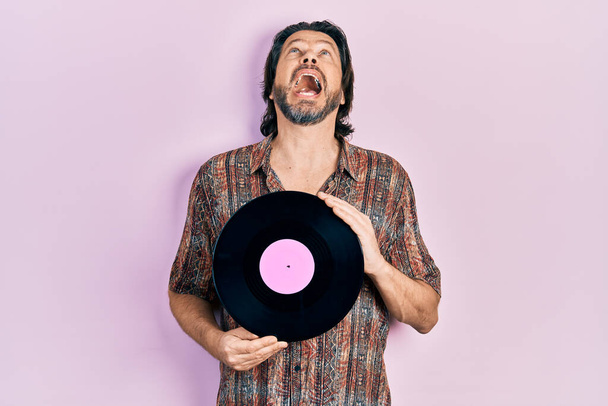 Middle age caucasian man holding vinyl disc angry and mad screaming frustrated and furious, shouting with anger looking up.  - Photo, Image