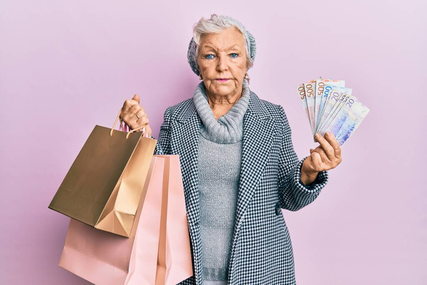 Idős ősz hajú nő bevásárlószatyrokkal és svéd koronabankjegyekkel lazítva, komoly arckifejezéssel. egyszerű és természetes nézi a kamerát.  - Fotó, kép