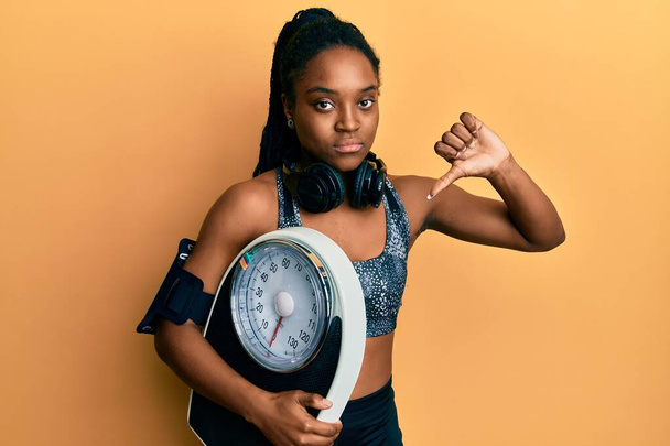 African american woman with braided hair wearing sportswear holding weighing machine with angry face, negative sign showing dislike with thumbs down, rejection concept  - 写真・画像