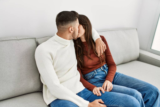 Молодая латиноамериканская пара сидит на диване, обнимаясь и целуясь дома. - Фото, изображение
