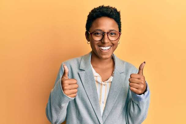 Giovane ragazza afroamericana indossa giacca da lavoro e occhiali segno di successo facendo gesto positivo con la mano, pollice in su sorridente e felice. espressione allegra e gesto vincente.  - Foto, immagini